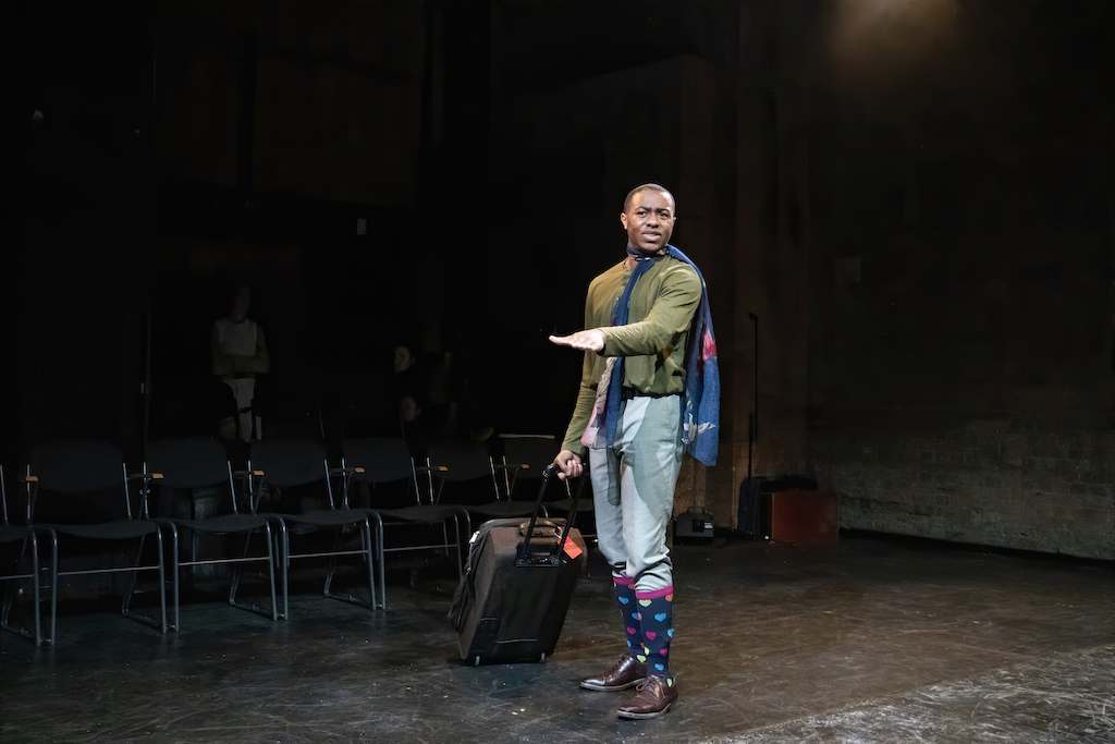 Actor wearing a green top, blue trousers, and heart-print knee socks pulls a rolling suitcase on stage in mid-performance.